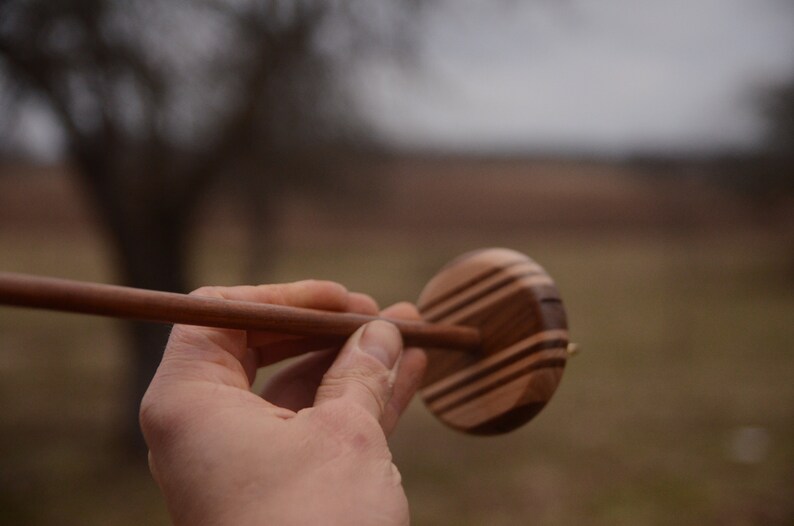 May include: A wooden spinning wheel with a brown and black striped design. The wheel is held in a person's hand.