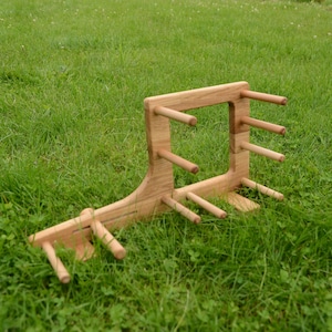 Op de afbeelding: Een houten garenwinder, een hulpmiddel om garen tot een streng te wikkelen. De winder is gemaakt van lichtgekleurd hout en heeft meerdere pinnen om het garen vast te houden. Het staat op een bed van groen gras.