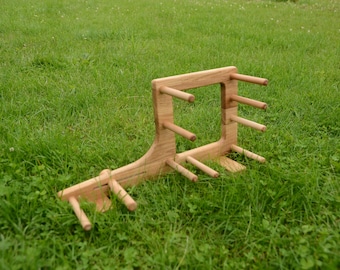 May include: A wooden yarn swift, a tool for winding yarn into a skein. The swift is made of light-colored wood and has multiple pegs to hold the yarn. It is set on a bed of green grass.