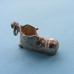 May include: A small, silver-colored boot charm with a textured surface. The charm has a loop at the top for attaching to a bracelet or necklace. The boot's design includes details like laces and a rounded toe.