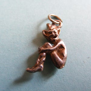 May include: A metallic silver-toned charm shaped like a small, seated figure with pointed ears and a smiling expression. The figure is depicted in a crouching position with knees pulled towards the chest, featuring a small circular jump ring at the top.