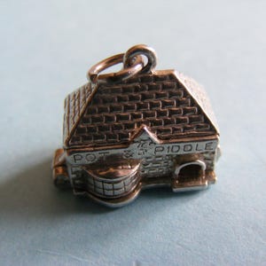 Könnte beinhalten: Ein silberfarbener Anhänger in Form eines kleinen Gebäudes, möglicherweise ein Pub oder ein Geschäft. Das Dach hat ein Ziegelmuster, und die Worte "POT & PIDOLE" sind sichtbar. Der Anhänger hat eine Schlaufe zum Befestigen.