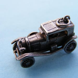 Op de afbeelding: Een gedetailleerde zilverkleurige miniatuur auto bedel. Het vintage voertuig heeft een klassiek ontwerp met zichtbare wielen, een dak en ingewikkelde details. De bedel is opgehangen aan een kleine lus, wat suggereert dat hij als hanger of decoratief element gebruikt kan worden.