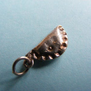 Könnte beinhalten: Ein kleiner, silberfarbener Anhänger in Form eines Halbkreises, der einem Pierogi oder einer Teigtasche ähnelt. Der Anhänger hat eine strukturierte Oberfläche und einen dekorativen Rand. Ein kleiner Silberring ist zum Aufhängen befestigt.