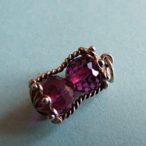 May include: A pendant featuring two faceted, deep purple beads encased in a silver-toned metal cage. The cage is constructed from twisted wire pillars and decorative filigree end caps, with a small circular jump ring attached to the top for hanging.