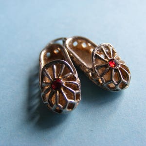 May include: A pair of silver-toned miniature shoe charms. Each shoe features a red gemstone in the center and an openwork design. The charms are displayed on a light blue background.