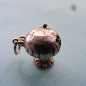 May include: A small, spherical, metallic charm with a tarnished silver-tone finish. The object features a hinged top, a small loop for attachment, and a textured base. A tiny, square-patterned cutout is visible on the side of the rounded body.