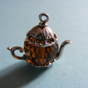 Könnte beinhalten: Ein Miniatur-Teekannen-Anhänger mit einem facettierten, bernsteinfarbenen Kristallkörper. Das Stück verfügt über einen silberfarbenen Metalldeckel mit dekorativen, gewellten Rändern, einen geschwungenen Griff, einen schmalen Ausguss und eine kleine Öse.