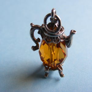 Peut inclure: Un charm en forme de théière argenté avec un corps en cristal facetté de couleur ambre. Le charm a une boucle pour l'accrocher et repose sur trois petits pieds. Le fond est bleu clair.