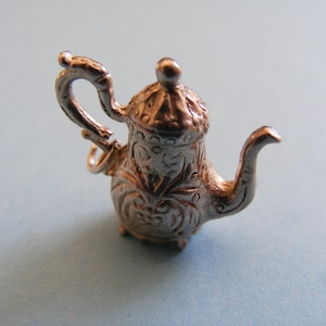 May include: A miniature silver-toned teapot charm featuring intricate embossed scrollwork patterns, a curved handle, a small spout, and a lid with a rounded finial. The metallic object includes a small circular jump ring attached to the handle.