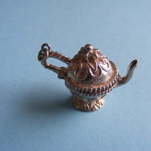 Könnte beinhalten: Ein Miniatur-Teekannen-Anhänger aus silberfarbenem Metall mit einem verzierten, geprägten Deckel mit blumenartigen Mustern, einem strukturierten Boden, einem geschwungenen Griff mit kleinem Ring und einem schmalen, nach oben gebogenen Ausguss.