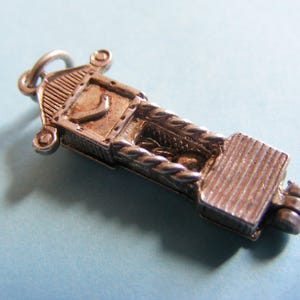 May include: A vintage silver-toned charm in the shape of a grandfather clock. The charm features detailed elements, including a clock face, a braided rope design, and a rectangular base. The charm is suspended from a small loop.