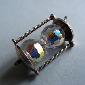 May include: A small, silver-toned hourglass pendant with a twisted frame. The hourglass contains two faceted, iridescent crystal beads. The pendant is likely a charm or a piece of jewelry.