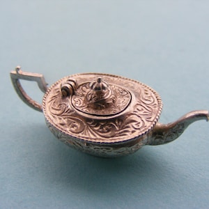 Puede incluir: Una pequeña tetera plateada con intrincados detalles florales y de volutas. La tetera presenta un pico curvo, un asa y una tapa decorativa con un pequeño remate. El diseño es vintage y ornamentado.