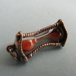 May include: A small, vintage hourglass charm with a clear glass center filled with orange sand. The metal frame has a twisted rope design and a loop for attaching to a bracelet or necklace. The charm is set against a light blue background.
