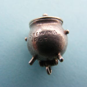 Könnte beinhalten: Ein kleiner, silberfarbener Anhänger in Form eines Kochtopfes. Auf der Vorderseite ist das Wort "STEWING SPOT" eingraviert. Der Topf hat einen Deckel, drei kleine Beine und zwei seitliche Griffe. Unten ist eine Schlaufe angebracht.