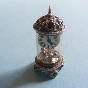 May include: A miniature silver-toned clock charm with a clear glass cylinder. The clock face is visible inside. The top and base have ornate detailing and red accents. A small loop is at the top for hanging.