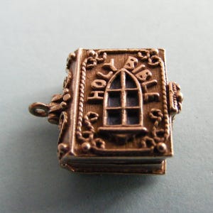 May include: A silver charm in the shape of a closed book with the words "Holy Bible" engraved on the cover. The charm has a detailed design with a window and ornate details.
