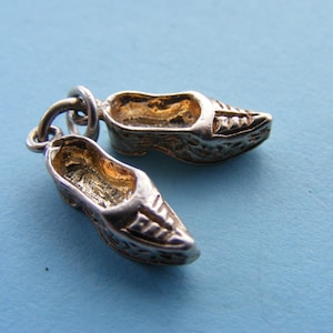 May include: A silver charm depicting two miniature clogs, connected by a small ring. The clogs have detailed textures and a gold-coloured interior, set against a light blue background.