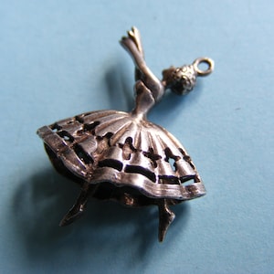 May include: A silver charm depicting a ballerina. The charm shows a dancer with arms raised, wearing a detailed tutu with cut-out patterns. It has a loop at the top for attaching to a bracelet or necklace. The charm is made of silver.
