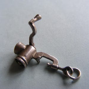 May include: A small, silver-toned charm in the shape of a miniature meat grinder. The charm features a handle, a crank, and a small loop for attachment. The design is detailed and appears to be vintage.