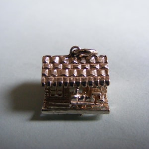 May include: A detailed, gold-colored charm in the shape of a small house. The charm features a textured roof and intricate detailing on the walls, with a loop at the top for attachment. The house charm is a miniature representation of a building.