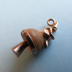 May include: A vintage charm depicting a figure on a mushroom-shaped base. The charm is crafted from gold-toned metal and wood, with a small loop for attachment. The mushroom has a silver-coloured band around the middle.