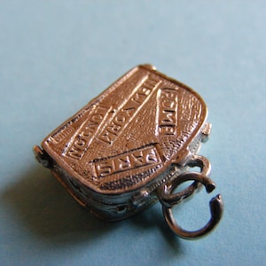 Puede incluir: Un charm plateado con forma de maleta. El charm está grabado con los nombres de las ciudades: París, Londres, Nueva York y Roma. Tiene un pequeño lazo para sujetarlo a una pulsera o collar.
