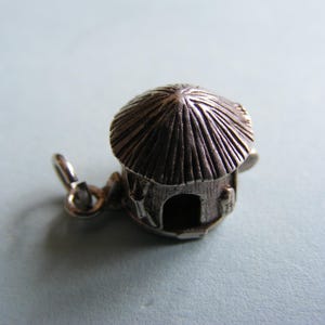 Op de afbeelding: Een zilverkleurige bedel in de vorm van een klein huisje met een rieten dak. Het huisje heeft een open deur en decoratieve details. De bedel heeft een lus om aan een armband of ketting te bevestigen. De bedel is gemaakt van metaal.