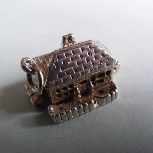 May include: A small, silver-colored house charm with a detailed roof, chimney, and windows. The charm has a loop for attaching to a bracelet or necklace. The house has a small fence around the base.