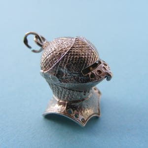 May include: A silver-toned metal charm shaped like a medieval knight's helmet. The helmet features a textured, cross-hatched surface pattern, a horizontal visor slit, and a flared neck guard. A small circular jump ring is attached to the top.