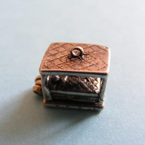 May include: A small, metallic, rectangular charm featuring a textured, cross-hatched roof design with a central jump ring for attachment. The structure has open sides and a detailed, textured interior base, finished in a dark, aged bronze tone.