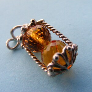 May include: A silver-toned pendant with two amber-colored, faceted beads encased in a decorative cage. The cage has a twisted rope design and ornate, scalloped edges. The pendant is on a light blue background.