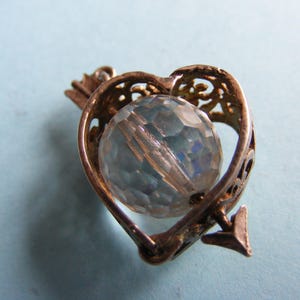 May include: A vintage-style pendant featuring a clear, faceted crystal orb encased within a heart-shaped frame. The frame is pierced with decorative filigree and an arrow design, suggesting a romantic theme. The metal has a tarnished, antique appearance.
