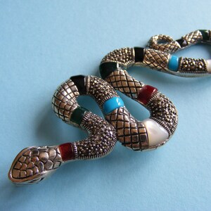 May include: A silver snake brooch with a textured, scale-like pattern. The brooch has black, green, red, and turquoise enamel accents, plus small, round, silver-coloured embellishments. A white, pearlescent stone is also incorporated into the design.