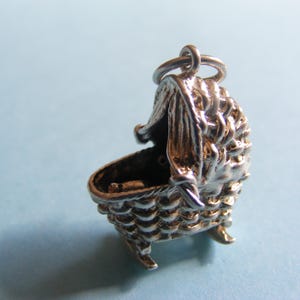 May include: A silver-colored charm in the shape of a baby cradle. The cradle has a textured, woven appearance with a hood and small feet. The charm is designed with a loop for attachment to a bracelet or necklace.
