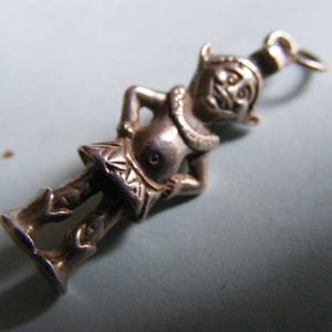 Puede incluir: Un charm de tono plateado con la forma de una figura caprichosa, posiblemente un duende o un diablillo. La figura tiene una cara sonriente, un collar decorativo y una prenda similar a una falda. El charm está diseñado con un lazo para sujetarlo a una pulsera o collar.