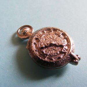 May include: A small, round, silver-toned metal charm shaped like a vintage pocket watch. The surface features intricate embossed scrollwork patterns, Roman numeral hour markers around the edge, and a small circular loop at the top for hanging.