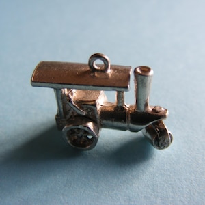 May include: A small, silver-colored train charm with a detailed design. The charm features a locomotive with a smokestack, wheels, and a small loop for attaching to a bracelet or necklace. The charm is set against a light blue background.