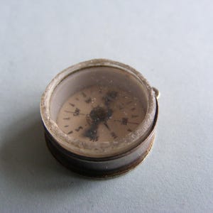 Pode incluir: Uma pequena bússola antiga com uma tampa transparente em forma de cúpula. A bússola tem uma caixa de metal prateado e um mostrador de cor clara com marcações pretas. A agulha aponta para o norte. Uma ferramenta de navegação vintage.