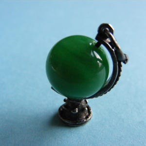 May include: A silver charm in the shape of a globe with a green glass orb. The globe is on a stand with a silver base.