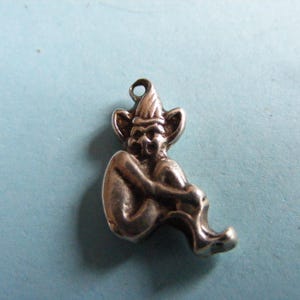 May include: A silver-toned charm depicting a seated goblin or elf figure. The charm has a pointed hat, large ears, and is curled up with its arms around its knees. The charm has a loop at the top for attaching to a necklace or bracelet.