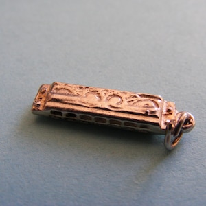 May include: A small, metallic harmonica charm featuring a detailed, embossed scrollwork pattern along the top surface. The rectangular instrument has a series of small, aligned air holes along the front edge and includes a circular jump ring attached.