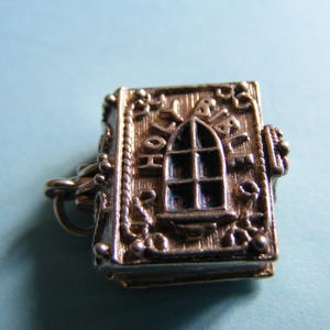 May include: A silver-toned charm in the shape of a book, featuring a window design on the front cover. The charm has intricate detailing, including decorative borders and what appears to be text. The background is a light blue.
