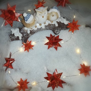 May include: A white owl and two birds with red, white, and blue hats are perched on a branch with a string of white lights and red paper stars. The scene is set on a white, snowy surface.