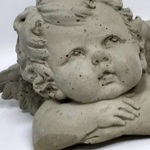May include: A gray concrete cherub statue with wings, the cherub is lying on its side with its head resting on its arm.