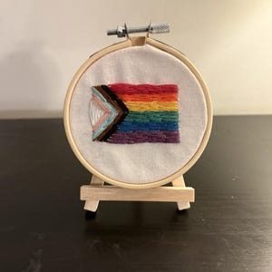 May include: Embroidered Pride flag in a wooden hoop, displayed on a small wooden easel. The flag features a rainbow design with additional colors representing inclusivity. The embroidery is detailed and colorful, showcasing the flag's vibrant hues.