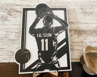May include: A black and white wooden basketball art piece featuring a player in a jersey with "OLSON" and the number "11". The player is depicted in the act of shooting a basketball. A basketball silhouette is also included.