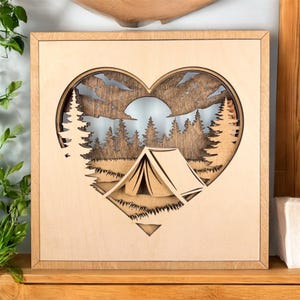 May include: Wooden wall art featuring a heart-shaped cutout with a camping scene. The design includes a tent, trees, and a sun, all crafted from layered wood. The frame is a light brown color, and the artwork evokes a nature theme.