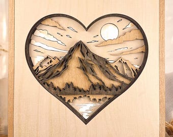 May include: A wooden wall art piece featuring a mountain landscape scene within a heart-shaped cutout. The design includes layered mountains, trees, clouds, and a sun, all in shades of brown and beige. The artwork is framed in a light-colored wood.